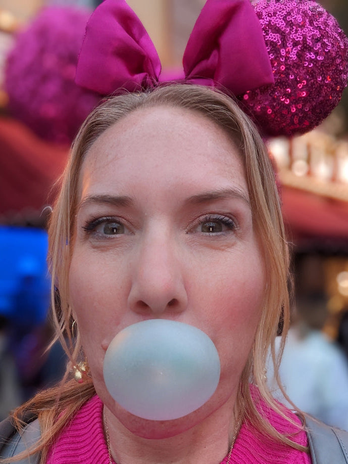 Person wearing pink Minnie Mouse ears blowing a bubble with pink bubble gum.